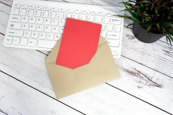Red note inside brown envelope with keyboard and potted plant background on wooden desk. Copy space. Flat lay.