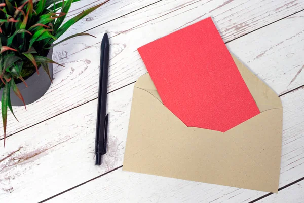 Red card mockup with envelope, pen and potted plant on wooden table. Copy space. Flat lay. Invitation concept.