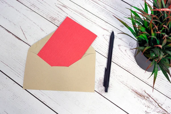 Red card mockup with envelope, pen and potted plant on wooden table. Copy space. Flat lay. Invitation concept.