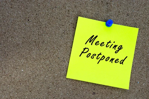 Meeting postponed on yellow stick note and pinned to a cork notice board. Reminder and business concept.