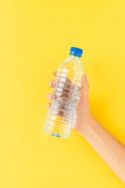 Sarı arka planda küçük plastik su şişesini tutan kadın.