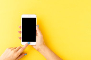 Sarı arka planda boş ekranı olan akıllı telefon kullanan kadın ellerinin yukarıdan çekimleri. Üst görünüm