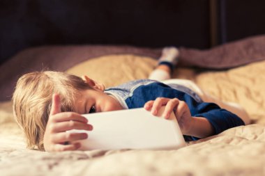 Smiling kid using digital tablet while lying down in the bedroom and looking at camera. 