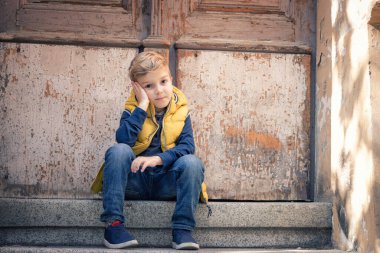 Small boy thinking while sitting in front of a door and waiting for someone. Bored child relaxing on stairs. 