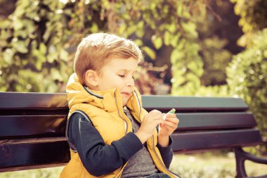 Small kid eating candy while relaxing on a bench in the park.