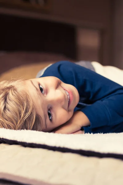 Happy little boy resting at home while lying down on bed and looking at camera. 