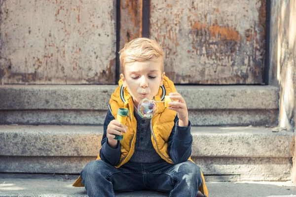 Small kid relaxing on steps and blowing soap bubbles.