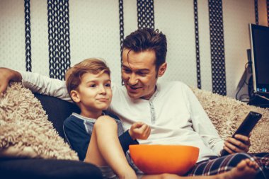 Happy kid and his father watching Tv while eating popcorn and relaxing in the living room. 