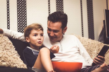Happy kid talking to his father while eating popcorn and watching TV together in the evening. 