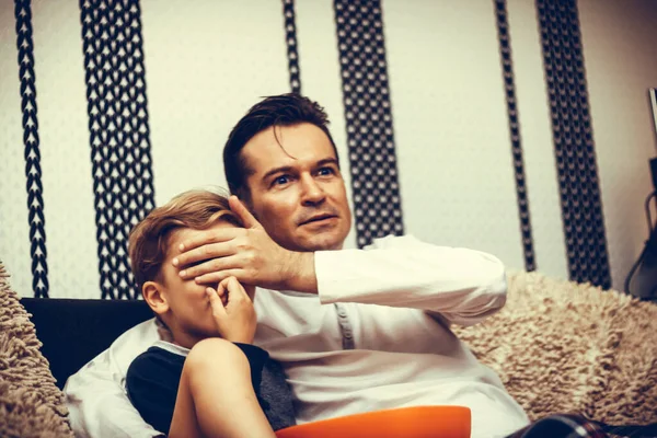 Father covering son\'s eyes while watching inappropriate television content at home.