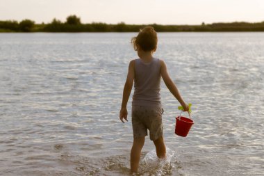 Rear view of kid with water bucket walking through the shallow water. 