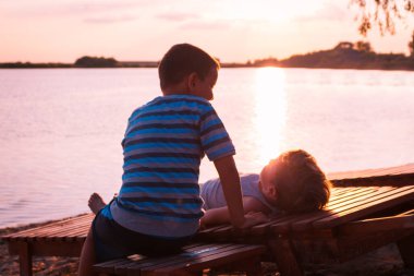 Two boys spending summer holiday at beach and relaxing on deck chairs at sunset.