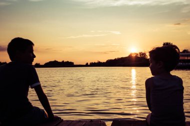 Small kid relaxing on a pier and enjoying in summer day by the water at sunset. 