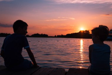 Two small boys sitting by the sea at sunset and enjoying in summer vacation. 