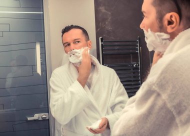 Reflection in a mirror of man applying shaving foam while shaving in the bathroom.