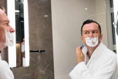 Man shaving his face with razor while looking himself in the mirror. 