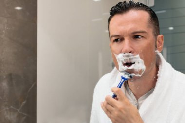 Man using razor and shaving his face while looking himself in the mirror. 