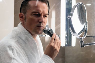 Man shaving beard with electric razor while looking himself in the mirror. The view is through the glass. 