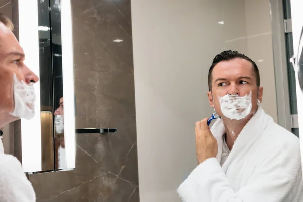 Man shaving his face with razor while looking himself in the mirror.