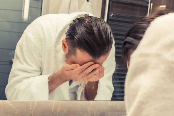 Reflection in a mirror of a man in bathrobe washing his face in the morning.