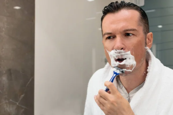 Man using razor and shaving his face while looking himself in the mirror.