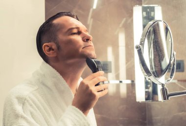 Man in bathrobe shaving with electric razor in the morning.