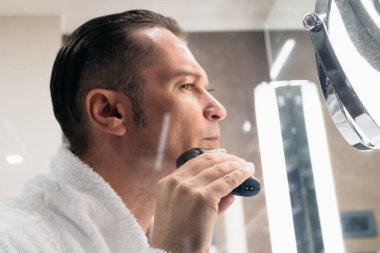 Close-up of man using electric razor while shaving in the bathroom. The view is through the glass. 