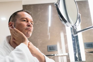 Man in bathrobe applying moisturizer after shaving his face in the morning. The view is through the glass.