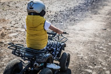 Back view of kid learning to drive four wheeled quad bike on dirt road.