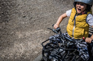 Little boy with mouth open driving four wheeled atv on dirt road. 