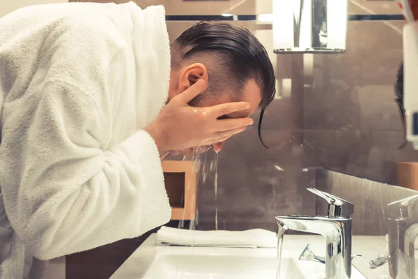 Side view of a man washing face in the bathroom sink.