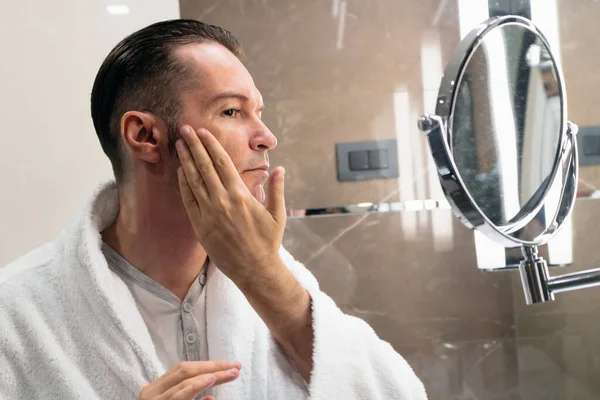 Man in bathrobe using face cream after shaving in the bathroom. The view is through the glass.