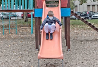 Playful kid with face mask having on a slide at the playground.  