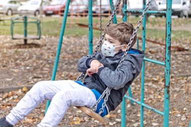 Lonely kid wit protective face mask swinging on the playground at the park. 