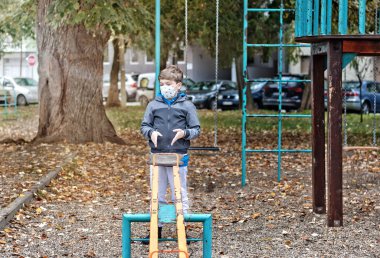 Little boy feeling bored while playing alone at the playground and wearing face mask due to COVID-19 pandemic.