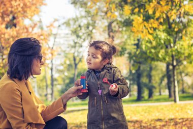 Mother assisting daughter while blowing soap bubbles in the park. Little girl talking to her mother while playing with bubble wand in nature.