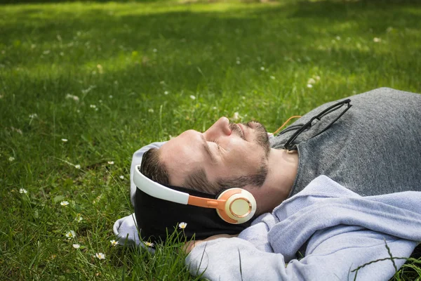 Carefree man lying on the grass and listening music over headphones with eyes closed in springtime.