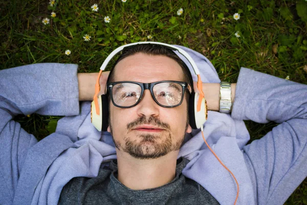 Close up of man lying on grass and listening music on headphones while daydreaming in spring day.