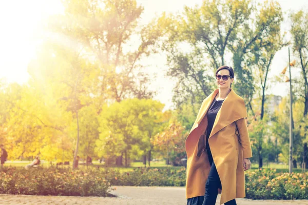 Beautiful fashionable woman walking in the park. Happy woman in a yellow coat enjoying in autumn day. 