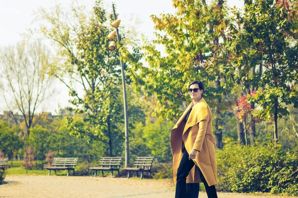 Fashionable woman walking in autumn park during sunny day. Beautiful confident woman taking a walk in city park.