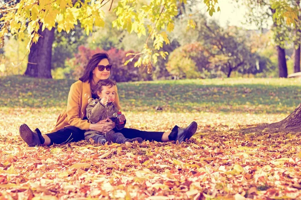 Smiling mother and daughter sitting on fall leaves and blowing bubbles in the park. 