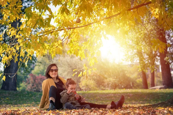 Happy mother enjoying with daughter in beautiful autumn day. Little girl blowing bubbles while sitting in autumn leaves at the park.