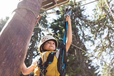 Below view of kid attaching carabiner on zip line while being on canopy tour in the forest. 