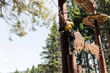 Small boy standing on a platform high up and attaching carabiner on zip line during canopy tour in the forest. 