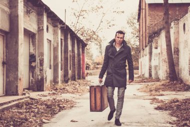 Serious man in coat walking thorough the street of a ruined city while carrying old leather suitcase.