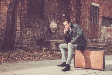 Worried man in a coat sitting on vintage suitcase and feeling disappointed while thinking about something.   