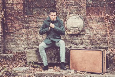 Fashionable man feeling cold and buttoning his coat while waiting on abandoned train station. Vintage leather suitcase in next to him. 