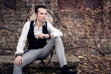 Portrait of handsome retro style gentleman sitting by brick wall and looking away. 