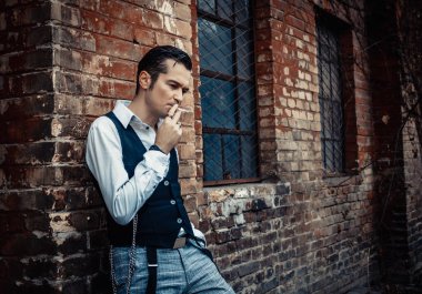 Serious retro0styled man smoking cigarette and thinking of something while leaning on a building outdoors. 