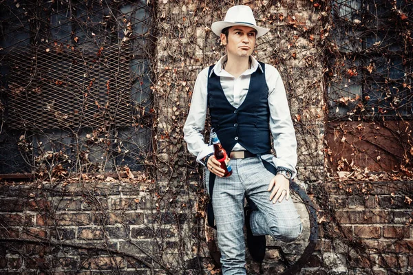 Retro-styled man with a hat thinking of something while holding beer bottle and leaning on brick wall.